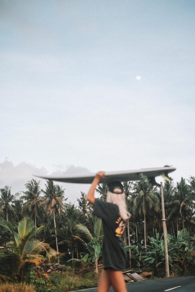 Remote-Workerin Leni Gruber mit Surfbrett vor einem Palmenwald