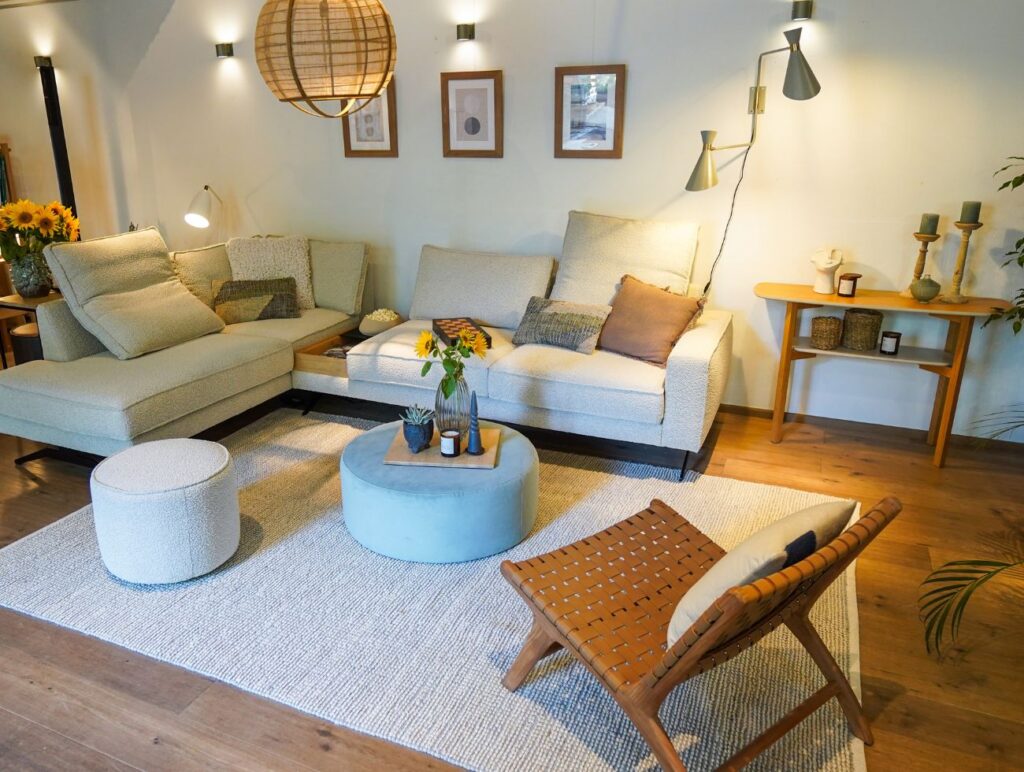 Das Wohnzimmer im Apartment zeuigt eine helle Eckcouch sowie ein geflochtener Loungesessel in Braun und runde Couchtische.