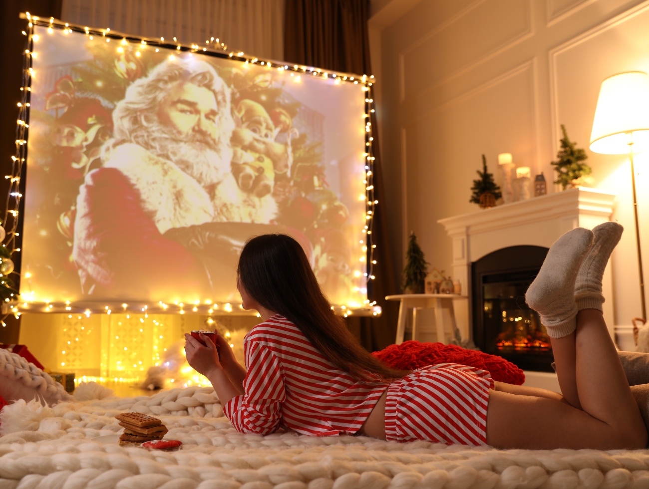Eine Frau liegt auf einer Kuscheldecke, trinkt Tee und sieht einen Weihnachtsfilm über einen Beamer an.