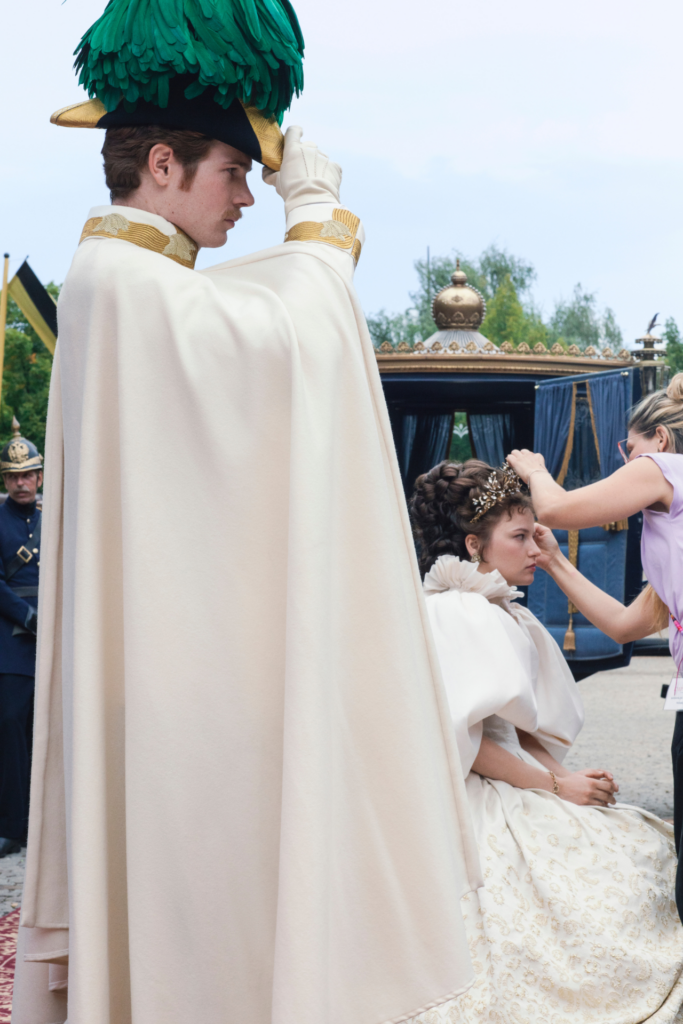 Die Kaiserin Staffel 2 auf Netflix (Behind the Scenes): Philip Froissant trägt einen Umhang und greift mit seiner Hand zu einem mit Federn besetzten Hut auf seinem Kopf. Devrim Lingnau knieht in ihrem Kostüm vor einer Kutsche, eine Stylistin fixiert ihre Krone im Haar.