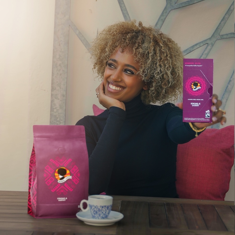 Neu: Afro Coffee Kapseln. Eine Frau hält die Packung hoch und lächelt dabei, vor ihr steht eine Kaffeetasse.