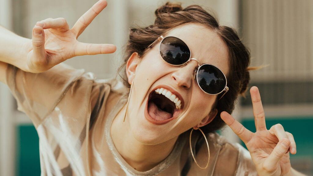 Challenge: Frau mit Sonnenbrille zeigt mit beiden Händen das Peace-Zeichen und lacht