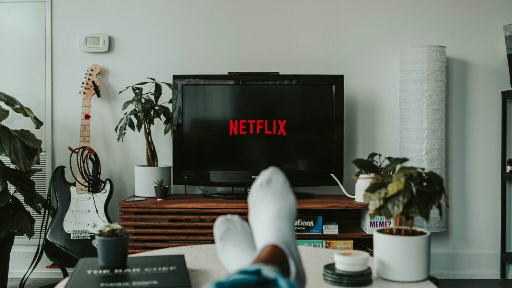 Serien: Aufnahme von Fernseher auf dem das Netflix Logo zu sehen ist. Man sieht Füße auf dem Couchtisch liegen. neben dem Fernsehern steht eine E-Gitarre.