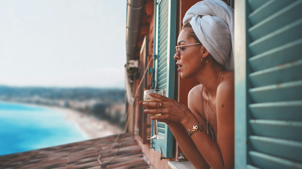 Solo-Trip: Junge Frau mit Handtuchturban, Sonnenbrille und Glas in der Hand schaut aus einem Fenster auf einen Strand