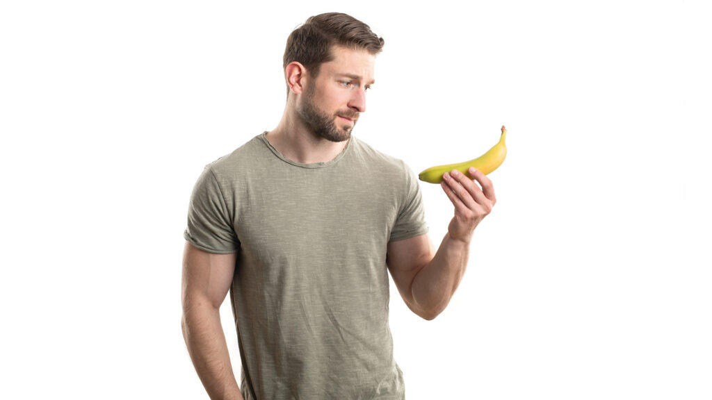 Mythos Penis: Attraktiver Mann mit Bart schaut skeptisch auf eine Banane