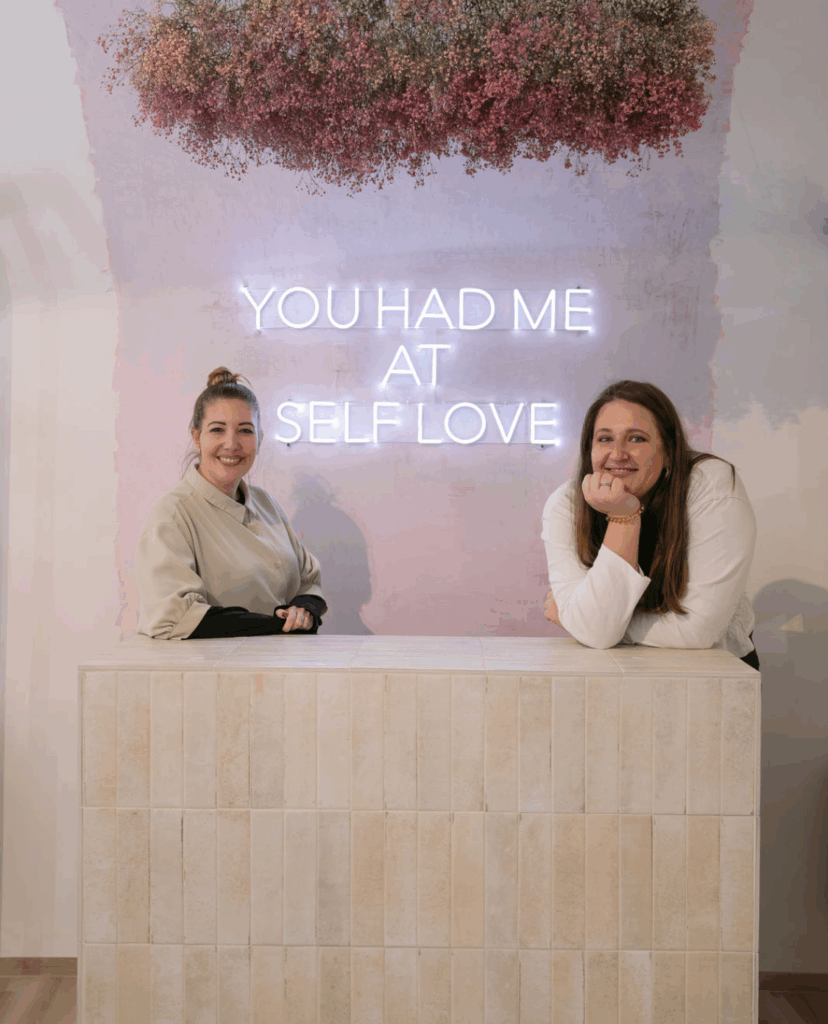 Die Inhaberinnen des Selfloveclub Salzburg: Caterina Müller und Julia Wenderoth im Porträtbild. Im Hintergrund "You had me at selflove" in Neonlichtern