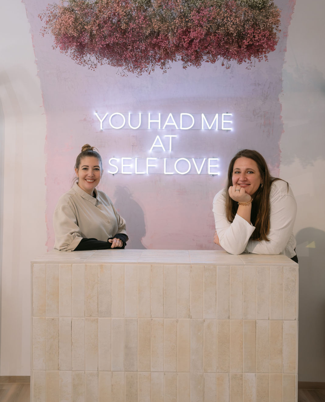 Die Inhaberinnen des Selfloveclub Salzburg: Caterina Müller und Julia Wenderoth im Porträtbild. Im Hintergrund "You had me at selflove" in Neonlichtern