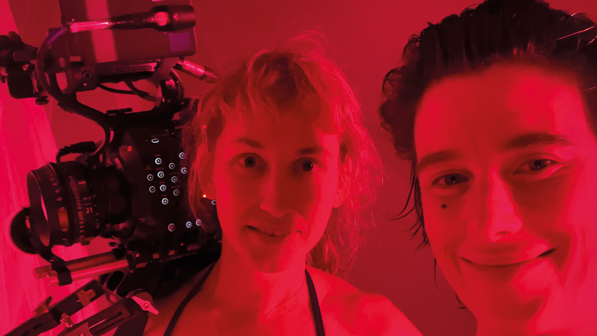 woman/mOther: Zwei Personen fotografieren sich neben einer Filmkamera in rotem Licht, beide blicken in die Kamera.
