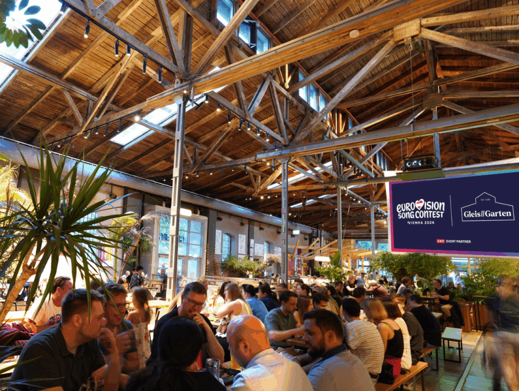 Menschen sitzen auf Bänken in einer Halle mit einem Bildschirm zur Übertragung des ESC