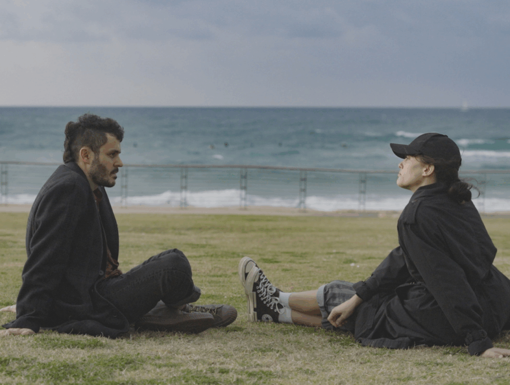 Filmszene aus "Noga", Noga Erez und ihr Partner Ori Rousso sitzen auf einer Wiese am Meer